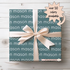 May include: A teal gift box with a white pattern of the words "In mason" and "ason" repeated. The box is tied with a cream-colored satin ribbon and bow. A speech bubble with a pink background and black outline says "over 50 colors available".