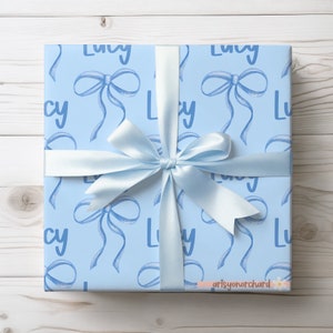 May include: A light blue gift wrap with a repeating pattern of the name "Lucy" and blue watercolor bows. The gift is wrapped and tied with a light blue ribbon.