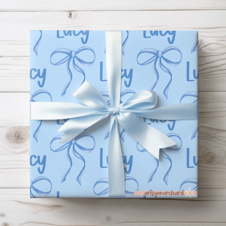 May include: A light blue gift wrap with a repeating pattern of the name "Lucy" and blue watercolor bows. The gift is wrapped and tied with a light blue ribbon.