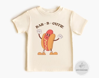 May include: A cream-coloured t-shirt with a cartoon hot dog character. The hot dog has a smiling face, raised arms, and shoes. The text "BAR-B-CUTIE" is arched above the hot dog. A fun, playful design for children.