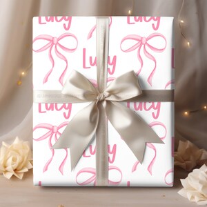 Peut inclure: Papier cadeau blanc avec un motif de n&oelig;uds roses et le nom "Lucy" r&eacute;p&eacute;t&eacute; partout. Le cadeau est emball&eacute; avec un ruban satin&eacute; beige.
