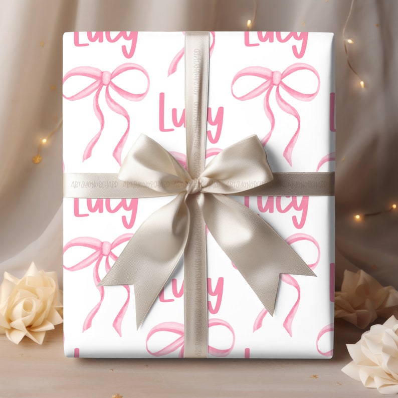 Peut inclure: Papier cadeau blanc avec un motif de n&oelig;uds roses et le nom "Lucy" r&eacute;p&eacute;t&eacute; partout. Le cadeau est emball&eacute; avec un ruban satin&eacute; beige.