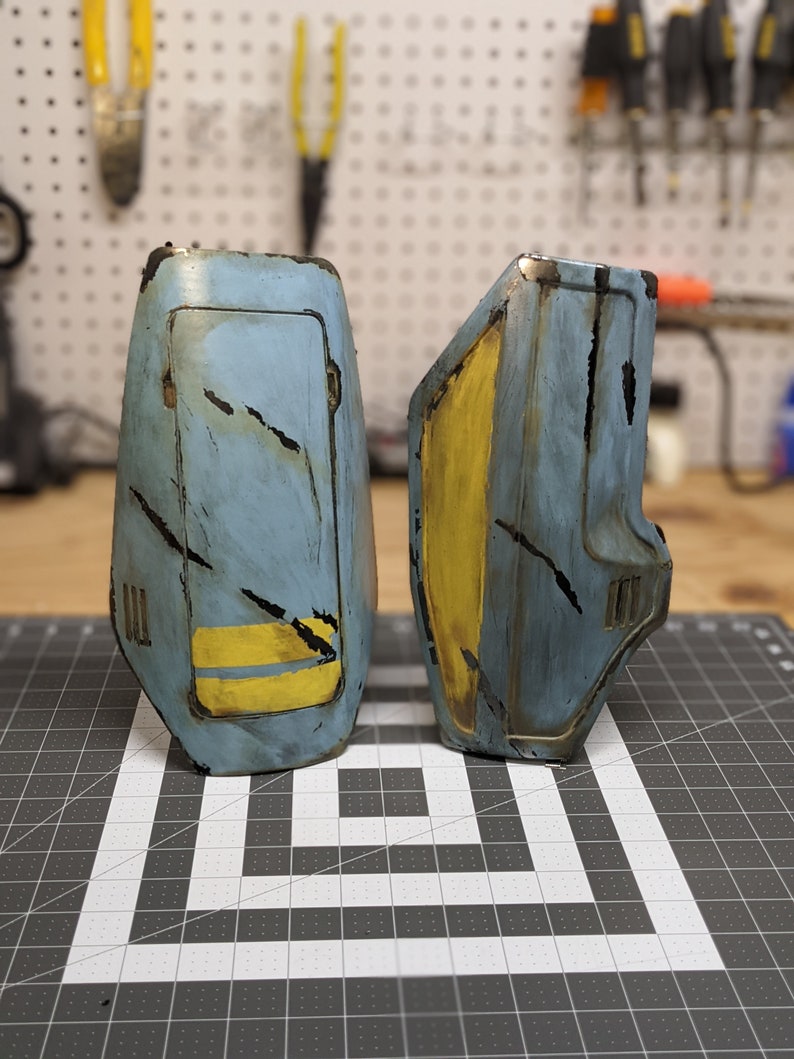 May include: Two blue and yellow painted metal pieces with a distressed look. The pieces have a rectangular shape with a yellow stripe and a small rectangular opening.