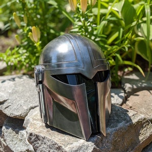 May include: A silver and green Mandalorian helmet with a black visor. The helmet is sitting on a pile of rocks.