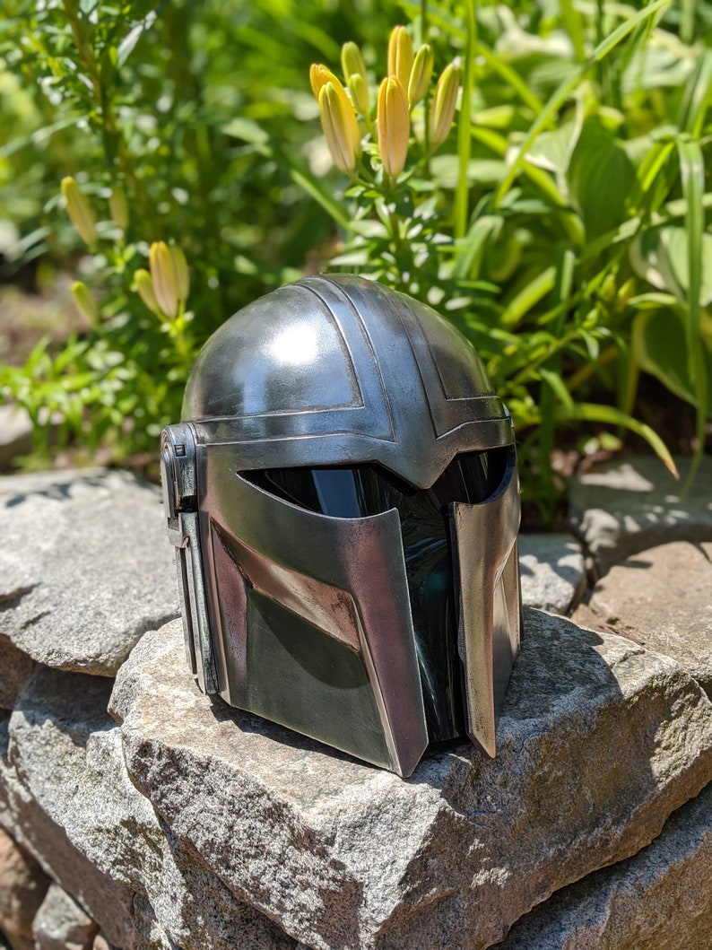 May include: A silver and green Mandalorian helmet with a black visor. The helmet is sitting on a pile of rocks.