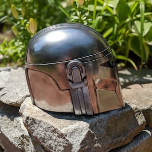 May include: A silver metal helmet with a textured surface and a raised detail on the side. The helmet is sitting on a pile of gray rocks.
