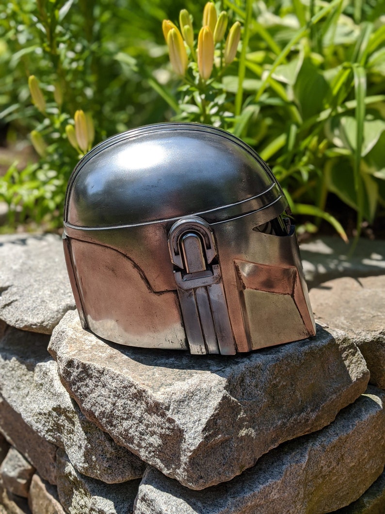 May include: A silver metal helmet with a textured surface and a raised detail on the side. The helmet is sitting on a pile of gray rocks.