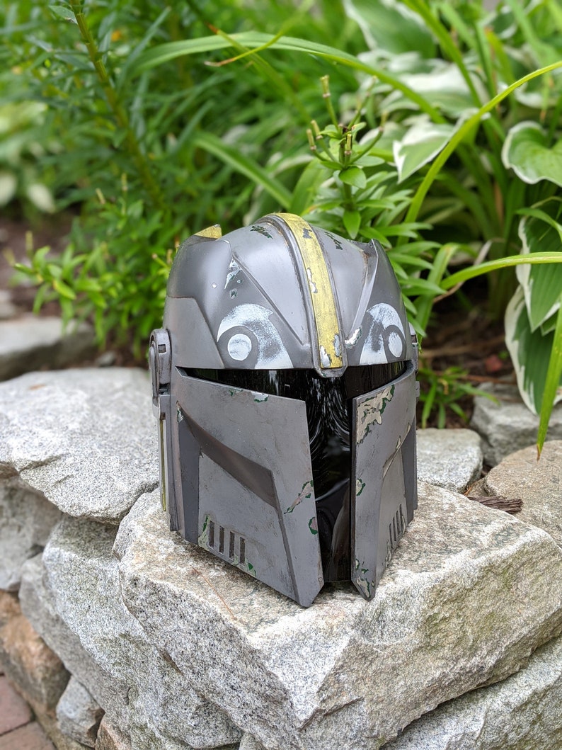 May include: A gray and yellow helmet with a weathered look. The helmet has a black visor and a design on the front.