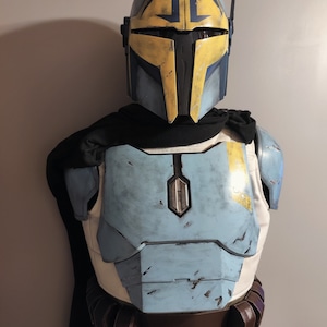 Puede incluir: Un casco mandaloriano azul y amarillo con una visera negra y una placa de pecho azul y amarilla con un aspecto desgastado.