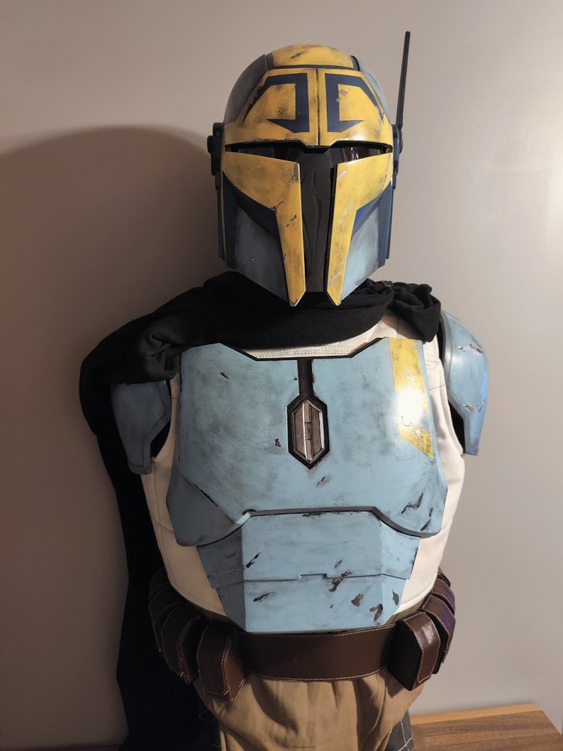 Puede incluir: Un casco mandaloriano azul y amarillo con una visera negra y una placa de pecho azul y amarilla con un aspecto desgastado.