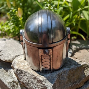 May include: A silver metal helmet with a textured surface and a raised design on the top. The helmet is sitting on a pile of gray rocks.