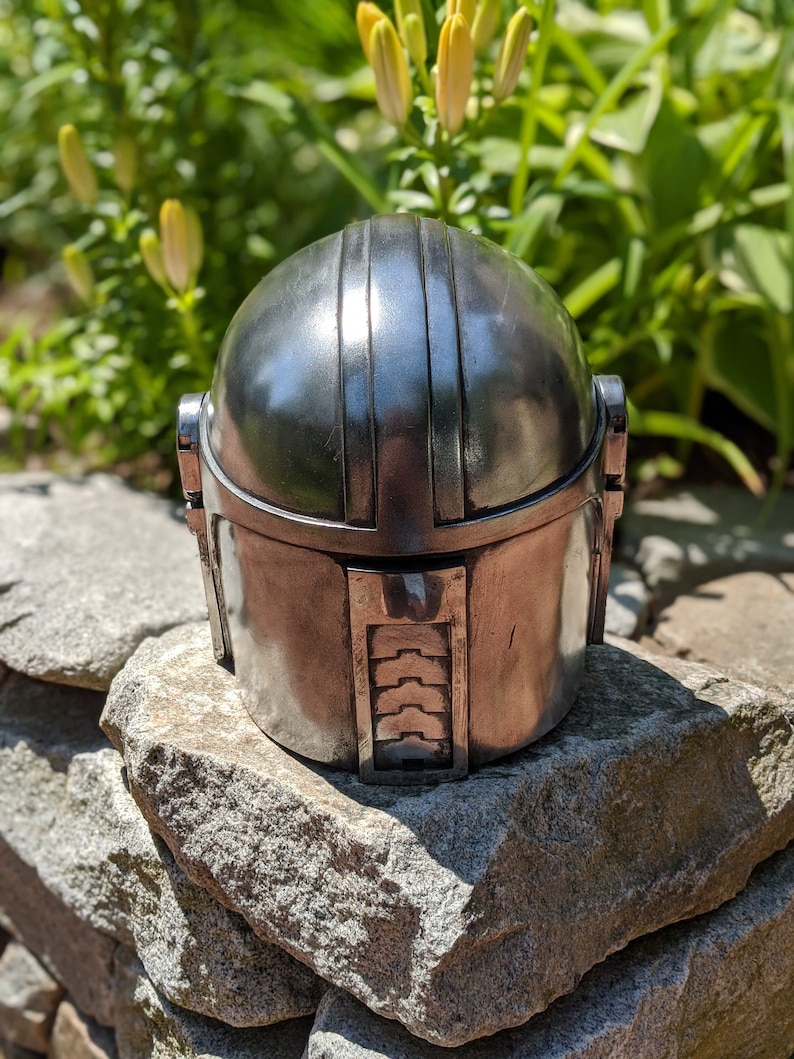 May include: A silver metal helmet with a textured surface and a raised design on the top. The helmet is sitting on a pile of gray rocks.