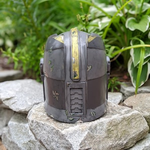 May include: A gray and yellow helmet with a weathered, distressed look. The helmet has a yellow stripe down the center and a series of vents on the side.