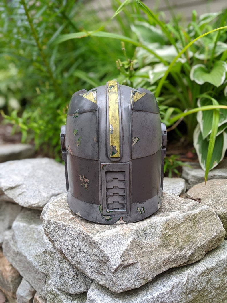 May include: A gray and yellow helmet with a weathered, distressed look. The helmet has a yellow stripe down the center and a series of vents on the side.