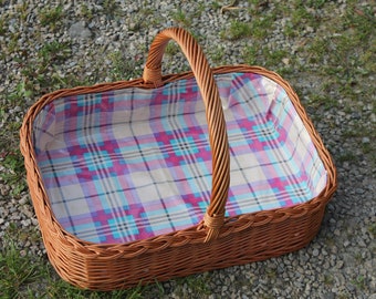 May include: A brown wicker basket with a plaid liner and a single handle. The basket is empty and sitting on a bed of gravel.