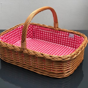 Könnte beinhalten: Ein rechteckiger Picknickkorb aus Weide mit einem Griff. Der Innenraum ist mit rot-weiß kariertem Stoff ausgekleidet. Der Korb besteht aus geflochtener brauner Weide und ist leer.