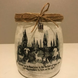May include: A white glass jar with a brown twine bow and a silver castle charm. The jar features a black and white silhouette of Hogwarts castle with a quote that reads "Happiness can be found even in the darkest of times, when one only remembers to turn on the light."