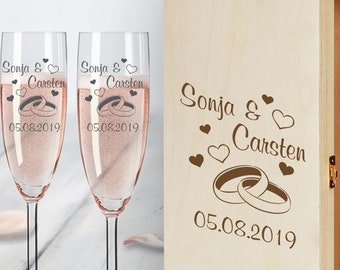 May include: Two champagne flutes and a wooden box with engraved text. The flutes are engraved with "Sonja & Carsten 05.08.2019" and two intertwined rings. The box is engraved with "Sonja & Carsten" and two intertwined rings with a heart above the text. The date "05.08.2019" is engraved below the rings.