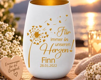 Puede incluir: Un vaso blanco con interior dorado que presenta un gráfico de diente de león dorado con semillas en forma de corazón y el texto en alemán "Für immer in unseren Herzen! Finn 28.01.2022" en dorado. Un pequeño corazón de madera con el texto "Erinnerungen für die Ewigkeit" está cerca.