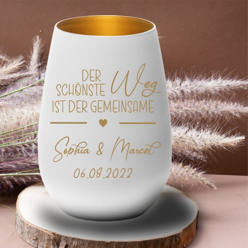 May include: White ceramic wine glass with a gold rim. The glass has the text "Der sch&ouml;nste Weg ist der gemeinsame" with a heart symbol and the names "Sophia & Marcel" and the date "06.08.2022" in gold lettering.