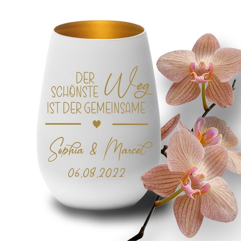 May include: White ceramic wine tumbler with a gold rim. The tumbler has a gold inscription that reads "Der sch&ouml;nste Weg ist der gemeinsame" with a heart symbol and the names "Sophia & Marcel" and the date "06.08.2022". The tumbler is sitting on a white surface with pink orchid flowers in the background.
