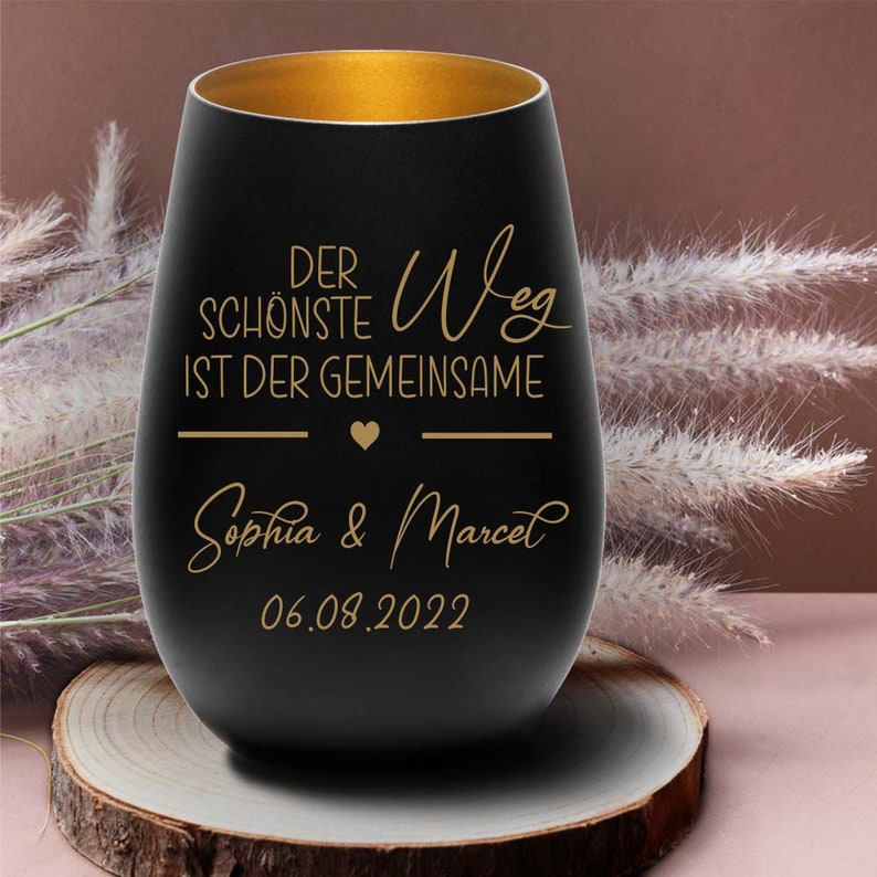 May include: Black glass candle holder with a gold rim. The candle holder has the text "Der sch&ouml;nste Weg ist der gemeinsame" with a heart symbol and the names "Sophia & Marcel" and the date "06.08.2022" in gold lettering.