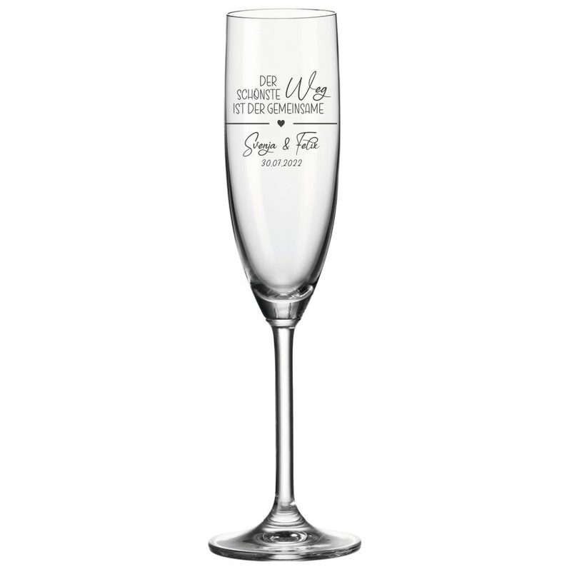 May include: A clear glass champagne flute with the text "Der sch&ouml;nste Weg ist der gemeinsame" and a heart symbol. Below the text is "Svenja & Felix" and the date "30.07.2022".