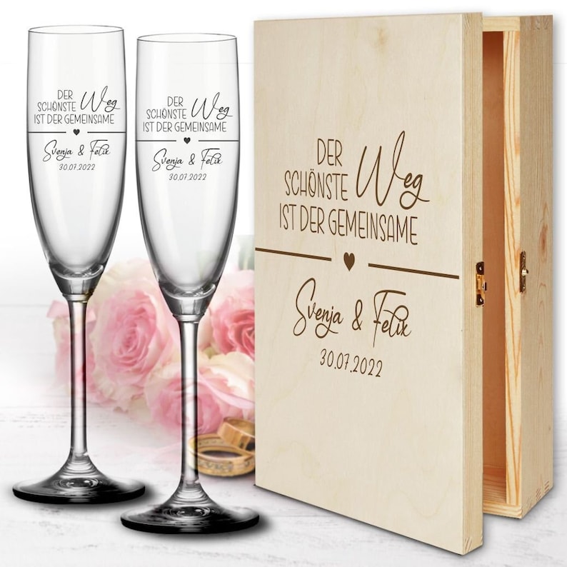 May include: Two clear glass champagne flutes with the text "Der sch&ouml;nste Weg ist der gemeinsame" and "Svenja & Felix 30.07.2022" engraved on them. The flutes are in a wooden box with the same text engraved on the front.