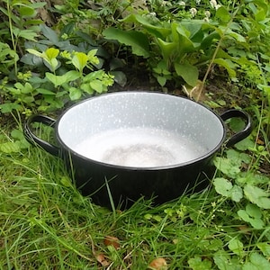 Könnte beinhalten: Ein schwarzer Emailletopf mit einem weißen, gesprenkelten Innenraum und zwei schwarzen Griffen. Der Topf steht auf einem Bett aus grünem Gras.