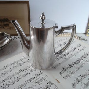 Könnte beinhalten: Eine silberne Vintage-Teekanne mit gebogenem Ausguss und Griff. Die Teekanne steht auf einem Blatt antiker Noten. Ein kleiner, dekorativer Knauf ziert den Deckel. Weitere silberne Servierteile sind im Hintergrund zu sehen.