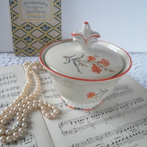 Könnte beinhalten: Eine Vintage-Zuckerdose aus Porzellan mit Deckel, mit einem floralen Design in Orange und Grau, akzentuiert mit einem roten Rand. Die Schüssel steht auf Notenblättern, mit einer Perlenkette und einem Buch im Hintergrund.