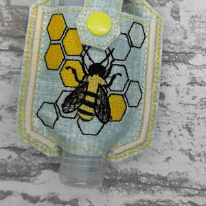 May include: A light blue fabric hand sanitiser holder with a yellow button closure. The holder features an embroidered bee and honeycomb design. The text "silly nana creations" is visible on the tag.