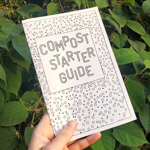May include: A white booklet titled "COMPOST STARTER GUIDE" held in a hand against a backdrop of green foliage. The cover features a dotted border and a rectangular box around the title.