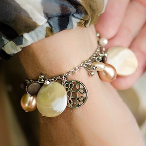 Op de afbeelding: Een zilveren bedelarmband met diverse hangers en bedels. De armband heeft ronde, ovale en druppelvormige bedels in crème-, perzik- en bruintinten. Eén bedel heeft een decoratief open ontwerp. De armband wordt om een pols gedragen.