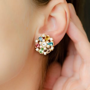 Puede incluir: Pendientes de botón dorados con diseño floral. Los pendientes presentan una piedra central transparente rodeada de piedras transparentes más pequeñas y gemas en forma de lágrima de colores en tonos azules, verdes, amarillos y rosas. Los pendientes están en la oreja de una persona.