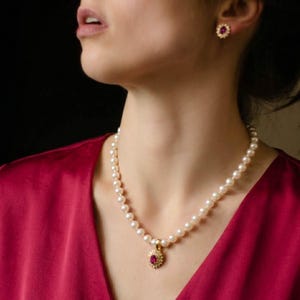 Könnte beinhalten: Eine Perlenkette mit einem Rubinanhänger und passenden Rubinohrringen. Der Anhänger und die Ohrringe sind von kleinen, funkelnden Steinen umgeben. Der Schmuck ist vor einem roten Hintergrund platziert.