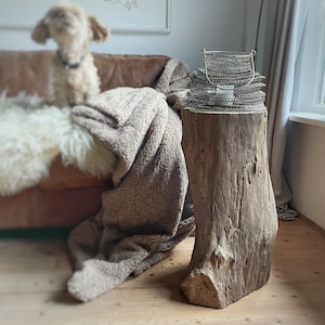 Könnte beinhalten: Ein rustikaler Beistelltisch aus Holz mit natürlicher, verwitterter Oberfläche. Auf dem Tisch befindet sich ein Stapel silberner Metallkörbe. Eine beige Decke und ein weißer Fellüberwurf sind über ein braunes Ledersofa drapiert. Im Hintergrund ist ein kleiner Hund zu sehen.