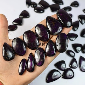 May include: A selection of polished, dark purple gemstones in diverse shapes, such as ovals, teardrops, and triangles. The stones have a glossy finish and show subtle colour variations. Some are held in a hand, while others are scattered on a white surface.