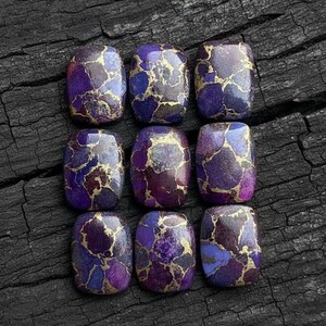 Puede incluir: Nueve piedras preciosas rectangulares con una mezcla de colores púrpura, azul y dorado. Las piedras tienen un aspecto veteado, con vetas doradas que atraviesan los tonos púrpura y azul. Las piedras están dispuestas en un patrón de cuadrícula.
