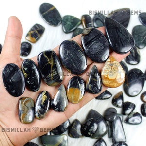 May include: A collection of black and gold gemstone cabochons, some with a teardrop shape, others oval, and some with a more irregular shape. The stones are arranged on a hand and a white surface. The text "Bismillah Gems" is visible in the image.
