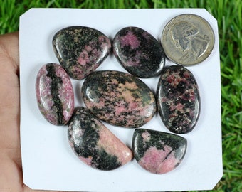 May include: A collection of polished gemstones in various shapes, including oval and teardrop. The stones feature a mix of pink, black, and green colors. A US quarter is included for size comparison. The gemstones are displayed on a white surface.