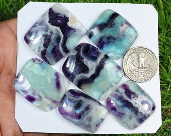 Può includere: Sette pietre di fluorite lucidata nei toni del viola, verde acqua e bianco, con una moneta per il confronto delle dimensioni. Le pietre sono rettangolari con angoli arrotondati, che mostrano motivi e venature uniche.