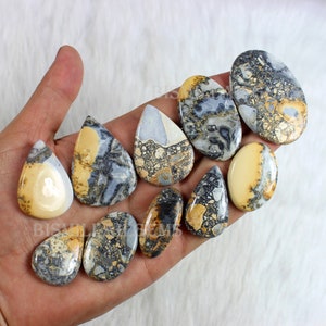 K&ouml;nnte beinhalten: Eine Sammlung von zehn tropfenf&ouml;rmigen, polierten, nat&uuml;rlichen Stein-Cabochons. Die Steine sind eine Mischung aus Creme, Braun und Grau mit einem marmorierten Muster.
