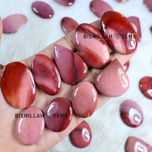 May include: A collection of polished red, pink and white gemstone cabochons. The cabochons are various shapes and sizes, including oval, teardrop and round. The cabochons are arranged on a hand, with the text "Bismillah Gems" visible on two of the cabochons.