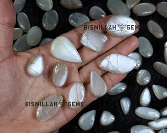 May include: A collection of polished, iridescent moonstone gemstones in various shapes, including oval, teardrop, and rectangular. The stones display a milky white and pale gray color with internal veining. The image includes the text "BISMILLAH GEM".