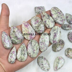 Peut inclure: Une collection de pierres précieuses polies de formes variées, dont des ovales et des gouttes. Les pierres présentent un mélange de teintes vertes, crème et roses, avec un aspect marbré. Le texte "BISMILLAH GEMS" est visible sur certaines pierres.