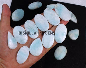 May include: A collection of 13 light blue aquamarine cabochons, some oval shaped and some teardrop shaped, on a hand. The cabochons are smooth and polished. BISMILLAH GEMS is printed on the hand.