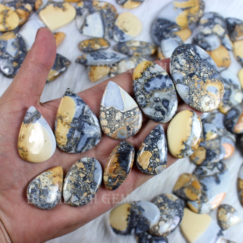 K&ouml;nnte beinhalten: Eine Sammlung von tropfenf&ouml;rmigen, polierten, nat&uuml;rlichen Edelstein-Cabochons. Die Steine sind eine Mischung aus Braun, Gelb, Grau und Wei&szlig; mit einem gesprenkelten Muster.