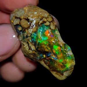Op de afbeelding: Een ruwe, ongeslepen opaal edelsteen met een levendige kleurenpracht. De opaal toont een mix van groene, oranje en blauwe tinten, gezet tegen een bruine matrix. De steen wordt vastgehouden, wat de natuurlijke, ruwe schoonheid benadrukt.