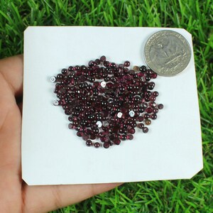 May include: A scattering of small, round, dark red gemstones, possibly garnets, on a white card. A UK quarter coin is placed nearby for size comparison. The gemstones are a deep, rich colour.